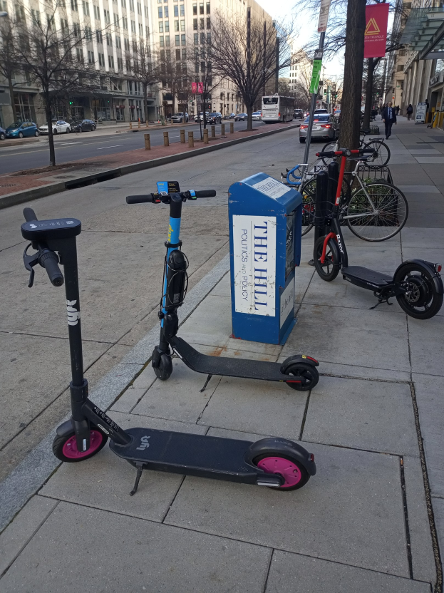 Zwei Elektro-Scooter auf einer Stadtstraße mit Fahrrädern, einem Müllcontainer und Fahrzeugen im Hintergrund geparkt.
