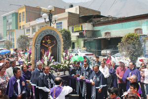 Menschen auf einer Straße vor der Stadt, mit einem Kreuz und Blumensträußen im Vordergrund, Autos in der Mitte und Gebäuden, Bäumen und Straßenlaternen im Hintergrund.
