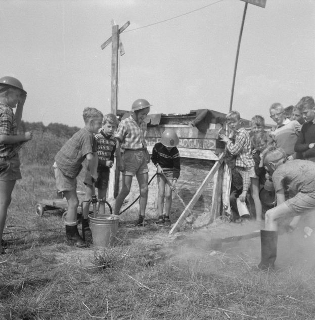 Schwarz-weißes Bild von Kindern in Helmen um ein Feuerhydrant in einem grünen Feld, mit einem Eimer, einer Karre, Stangen mit Drähten und einer Texttafel in der Nähe.