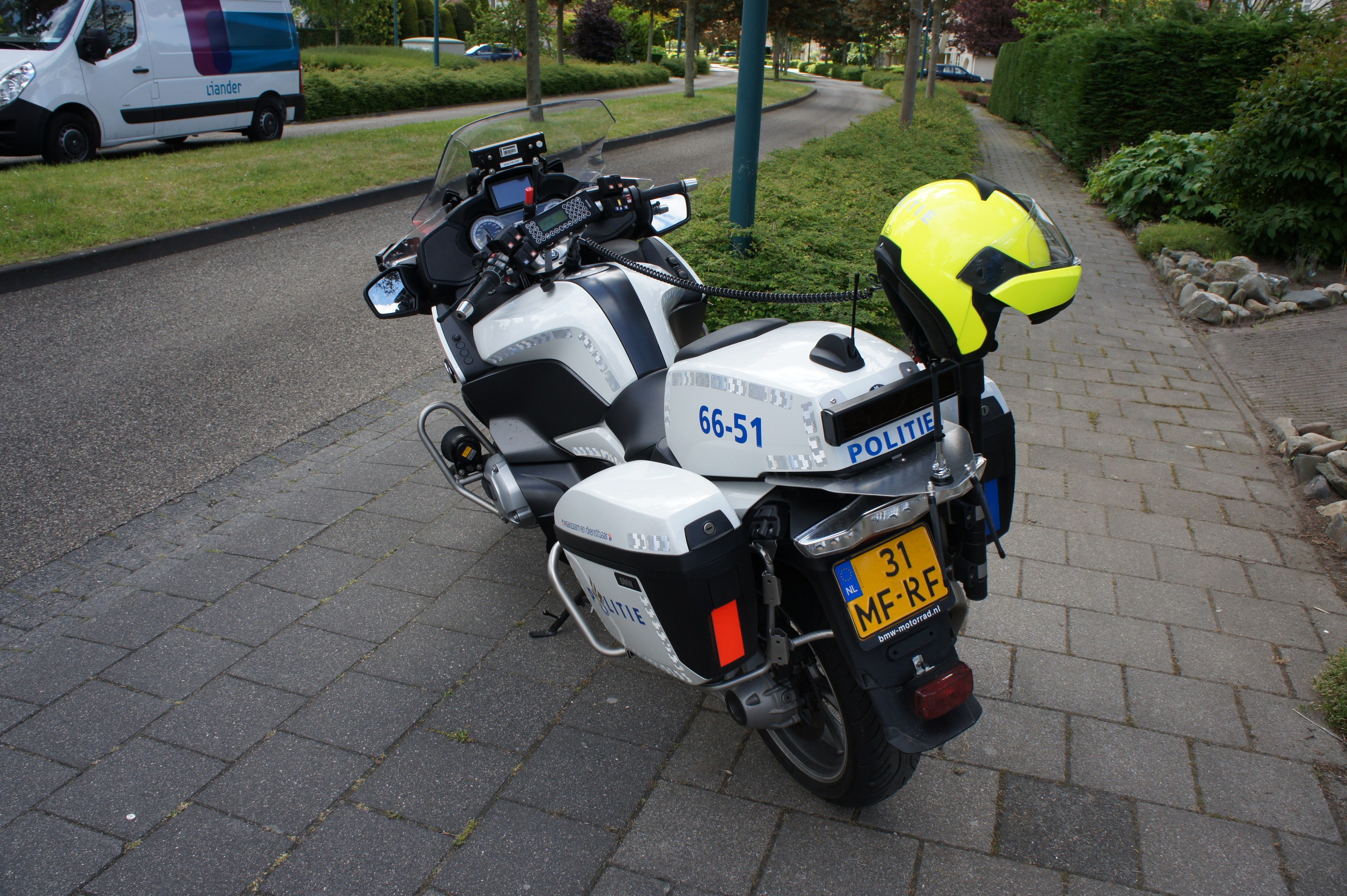Ein Polizeimotorrad am Straßenrand geparkt mit einem Helm darauf, umgeben von Bäumen, Gebäuden und anderen Fahrzeugen.