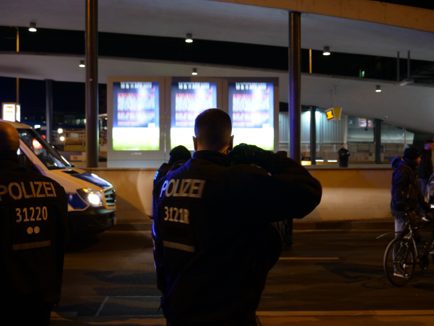 Eine Gruppe von Polizeibeamten steht vor einem hell erleuchteten Gebäude in der Nacht, mit einem geparkten Auto links und einem Radfahrer rechts.