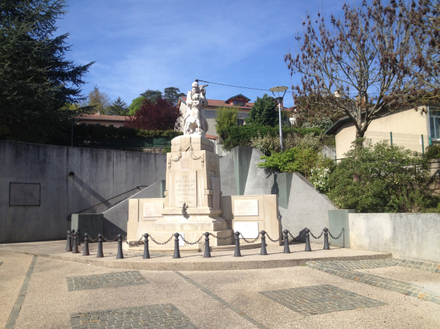 Eine bronzene Statue eines Mannes auf einem Sockel steht als Denkmal für die Pariser Anschläge, umgeben von einem Metallzaun, Bäumen, Pflanzen in einem Park mit Gebäuden und einem bewölkten Himmel.