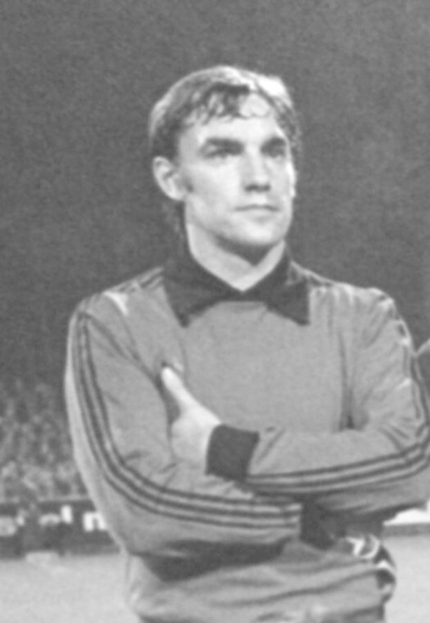 Schwarzes und weißes Foto eines Mannes, der mit verschränkten Armen auf einem Fußballfeld steht und von einer Gruppe von Sitzenden im Hintergrund umgeben ist.