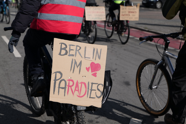 Eine Gruppe von Menschen, die auf Fahrrädern eine Straße entlangfährt, mit einem "Berlin I'm Paradies"-Schild im Vordergrund und einem Auto im Hintergrund, Bild etwas unscharf.
