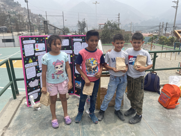 Gruppe von Kindern auf einem Basketballfeld, die Papiertüten halten, mit Tüten und Gegenst├Ąnden rechts und einer Tafel mit Text links, vor einem Hintergrund aus Z├Ąunung, Masten, Stra├čenger├Ąten, B├Ąumen, Geb├Ąuden, H├Ąngen und Himmel.