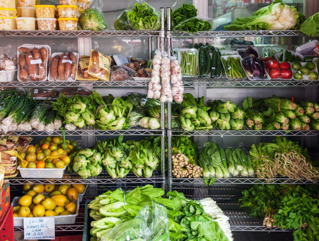 Ein Gang in einem Supermarkt mit frischem Gemüse und Obst in Plastikverpackungen, Kisten und einer Tafel im Hintergrund und einem Glasfenster weiter hinten.
