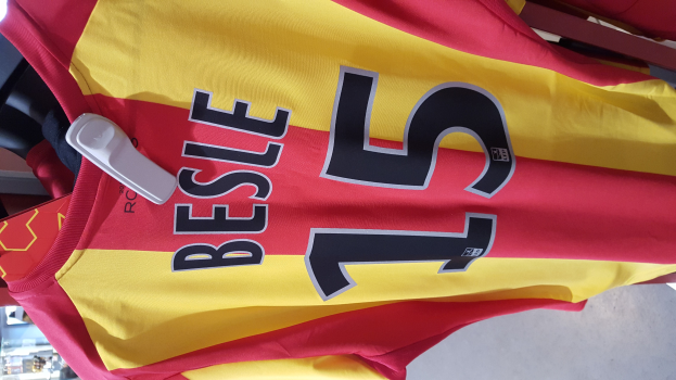 Ein rotes und gelbes Fußballtrikot mit aufgedrucktem Text und Nummern, das an einer Ladengestell hängend mit anderen Gegenständen und dem Hintergrund im Hintergrund zu sehen ist.