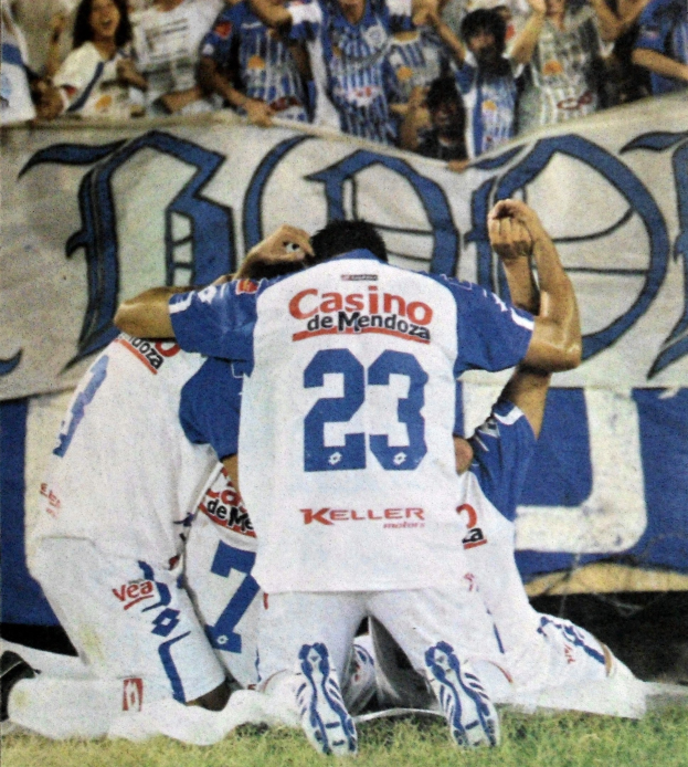 Gruppe von Fußballspielern feiert auf dem Boden mit jubelnden Zuschauern und einem Banner im Hintergrund.