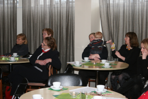 In einem Restaurant sitzen drei Frauen rechts auf Stühlen, drei Frauen links auf Stühlen und ein Tisch in der Mitte hält vier Tassen und Untertassen mit anderen Gegenständen, mit einer Säule und Vorhängen im Hintergrund.