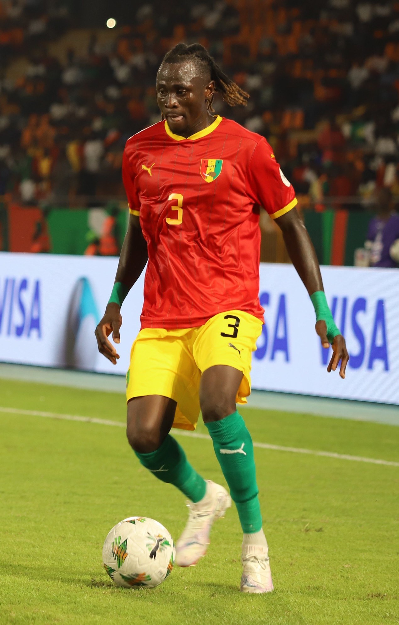 Ein Fußballspieler in rot-gelber Uniform, der einen Ball auf einem Feld kickt, mit Zuschauern auf den Rängen und Werbetafeln im Hintergrund.