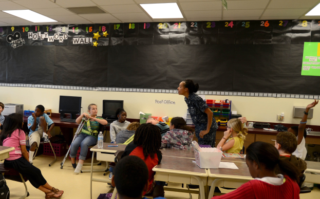 Ein Klassenzimmer mit Kindern an Tischen mit Monitoren, Flaschen, Körben und Papieren, während ein Lehrer in der Mitte unterrichtet.