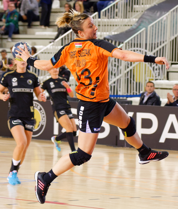 Eine Frau in einem orangen T-Shirt und schwarzen Shorts, die Handball spielt und einen Ball in der Hand hält, mit Menschen auf Treppen und Geländern im Hintergrund und einem Banner mit der Aufschrift "Krp Slezak - Handball-Weltmeisterschaft 2019".