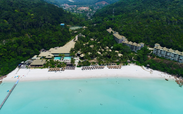 Luftaufnahme eines Resorts am Strand von Koh Samui, mit einer Brücke links, Gebäuden, Bäumen, Hügeln und einigen Menschen in Strandnähe.