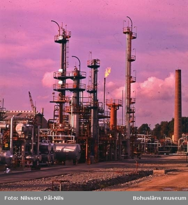 Ölraffinerie mit ausgedehnten Rohrleitungen, Metallkonstruktionen, Containern, Bäumen, einem Gebäude, einem Kran und einem bewölkten Himmel, mit Text unten.