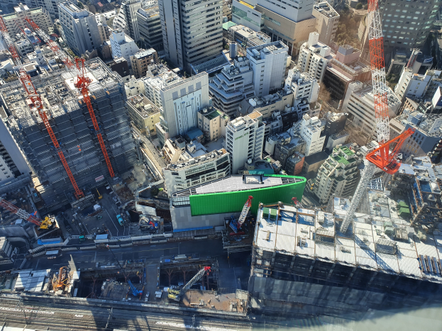 Eine Luftaufnahme einer belebten Stadt mit hohen Gebäuden, Kränen, Straße, Fahrzeugen und Bäumen, mit einer großeen grünen Baustelle in der Mitte.