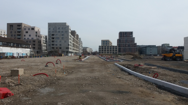 Baustelle mit schwerem Gerät im Vordergrund, Gebäude und Bäume im Hintergrund unter wolkenlosem Himmel.