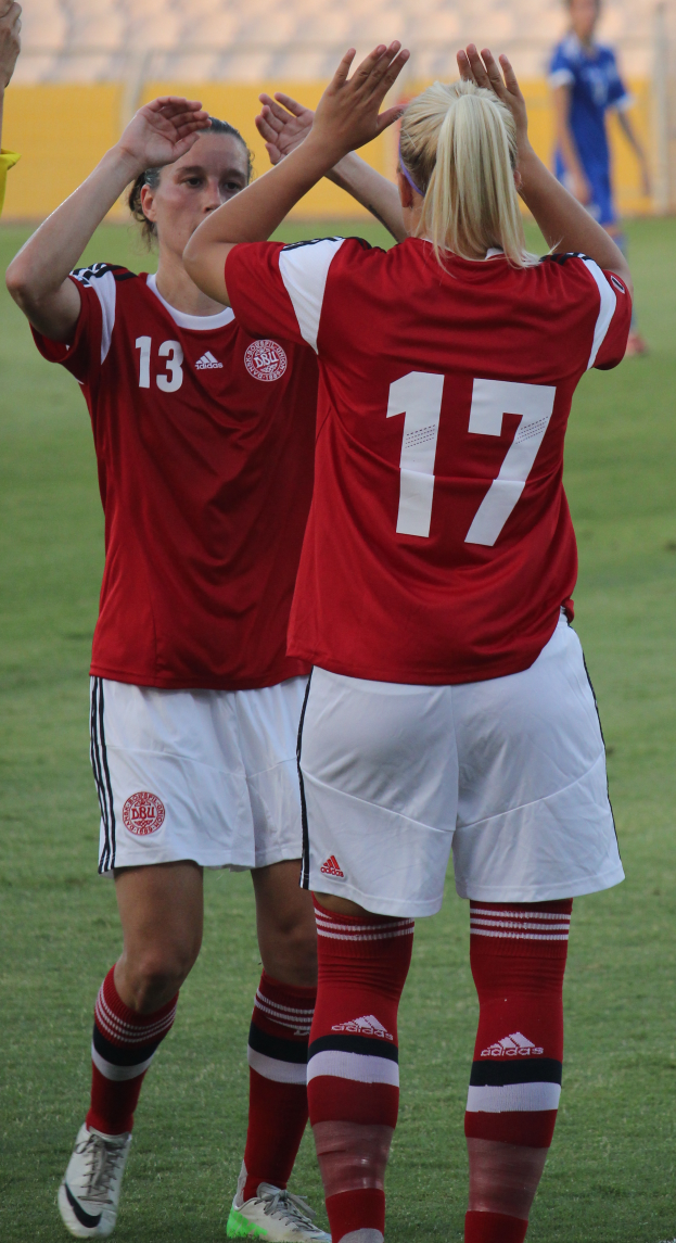 Zwei Frauen in roten T-Shirts und weißen Shorts stehen auf einem Fußballfeld und klatschen in die Hände, mit einer Person in einem blauen Kleid, einer gelben Tafel und ein paar Stühlen im Hintergrund.