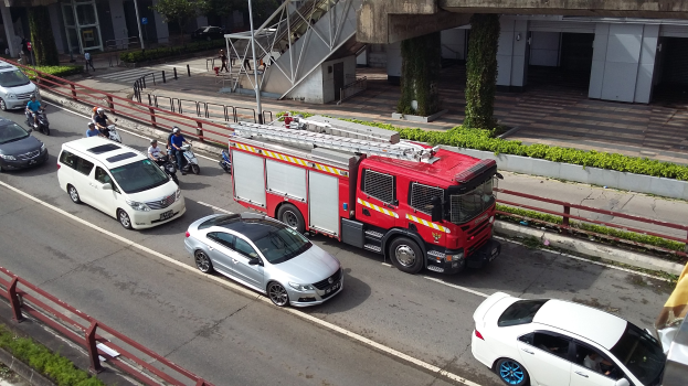 Feuerwehrfahrzeug fährt auf einer Straße mit Motorrädern, Autos und anderen Fahrzeugen; Gebäude, Bäume und Geländer sind im Hintergrund sichtbar.