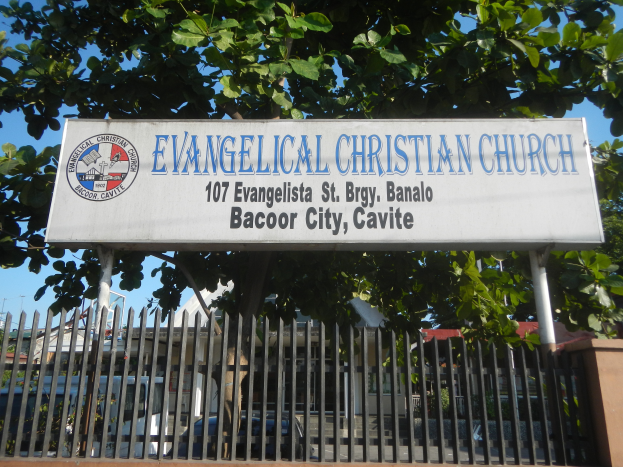 Eine evangelische Kirche in der Stadt Bacoor, Cavite, mit einem Schild, einem Metallzaun, einer Wand, Bäumen, Fahrzeugen, Gebäuden und Wolken am Himmel.