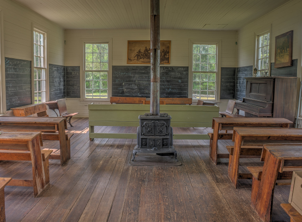 Leerer Klassenraum mit hölzernen Tischen und Stühlen in Reihen, eine mittlere Säule, ein Piano auf der rechten Seite und Wände mit Fenstern, Tafeln und Bäumen im Freien.