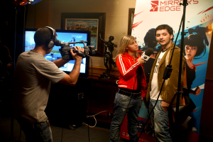 Ein Mann mit einer Videokamera und Headset filmt ein Interview zwischen einem Mann und einer Frau, mit einem Fernseher und einem gerahmten Foto im Hintergrund.