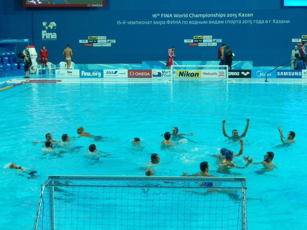Gruppe von Menschen, die Wasserball in einem Pool mit Netz am Boden spielen, Zuschauern und Informationsschildern im Hintergrund, einschließlich der Aufschrift "FINA World Championships 2015 Kazan".