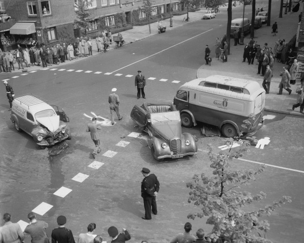 Schwarzes und weißes Bild eines Autounfalls an einer Straßenkreuzung mit mehreren Fahrzeugen, darunter ein Van, und Zuschauern; Gebäude, Bäume, Strommasten und Texttafeln im Hintergrund.