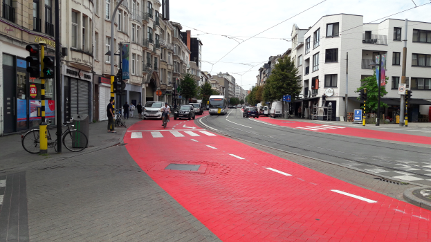 Eine Stadtstraße mit einem roten Radweg, Fahrzeuge auf der Straße, Fußgänger auf dem Gehweg, Gebäude mit Fenstern auf beiden Seiten und Bäume mit einem klaren blauen Himmel im Hintergrund.