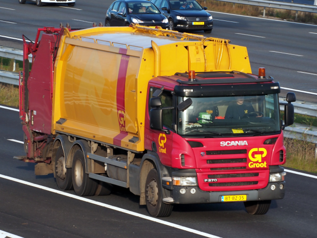 Ein gelber und roter Müllwagen mit einer Begrenzung fährt auf einer Autobahn, umgeben von anderen Fahrzeugen, mit Gras auf beiden Seiten der Straße.