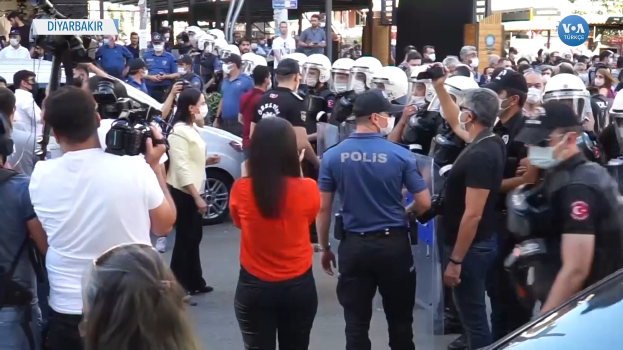Eine Gruppe von Menschen vor einer Reihe von Polizisten in Helmen, einige mit Kameras, mit Fahrzeugen, Gebäuden und Schildern mit der Aufschrift "Türkische Polizei im Konflikt mit Demonstranten in Ankara" im Hintergrund.
