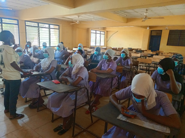 Ein Klassenzimmer in Nigeria mit Schülern, die Gesichtsmasken tragen und auf Bänken sitzen oder stehen, mit Büchern und Gegenständen auf den Tischen, Fenstern, einer Tür und Deckenleuchten und -ventilatoren im Hintergrund.