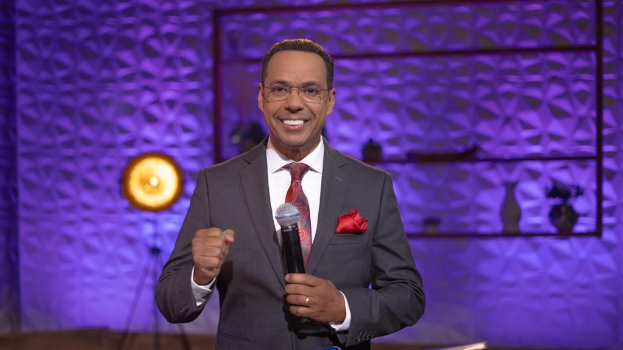 Stephen Colbert, Moderator von *The Daily Show with Trevor Noah*, steht auf einer Bühne und hält ein Mikrofon, trägt einen Anzug, eine Krawatte und eine Brille, mit einer Lampe und einem Ständer im Hintergrund.