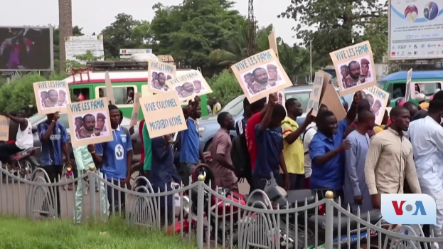 Eine Gruppe von Menschen hält Schilder, wahrscheinlich protestierend, vor einem Zaun mit Fahrzeugen, Bäumen, Masten, Absperrungen und einem Bildschirm im Hintergrund, unter einem sichtbaren Himmel mit Gras am unteren Bildrand.