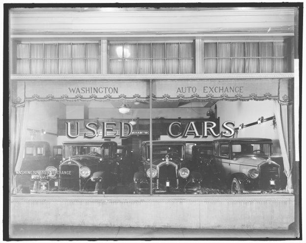 Schwarz-weiß-Foto eines Gebrauchtwagenhändlers in Washington, DC, mit mehreren Autos durch die Glasfront sichtbar und von Innenbeleuchtung erhellt.
