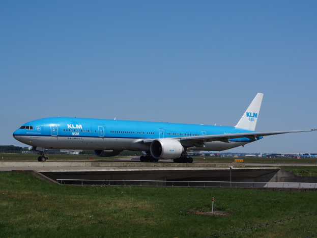 Ein KLM Boeing 777-300ER auf der Rollbahn am Amsterdam Airport, umgeben von Gras, einem Zaun mit Pfählen, Gebäuden, Bäumen und einem klaren blauen Himmel.