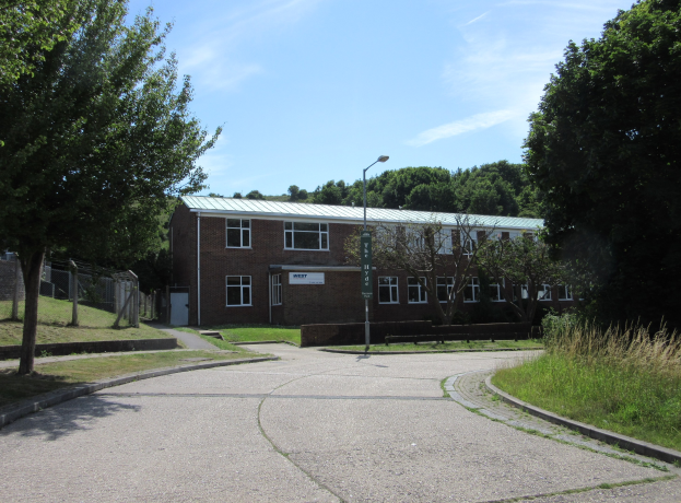 Ein Schulgebäude mit Fenstern, einem Straßenmast, einem Schild, einer Baumgruppe, Gras, einem Zaun und einem bewölkten Himmel, das inmitten eines Parks umgeben von saftigem Grün liegt.