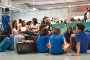 Ein belebter Flughafen-Terminal mit vielen Menschen, die auf ihre Flüge warten, Gepäckstücke auf der linken Seite und Glasdoors mit Richtungsschildern im Hintergrund.