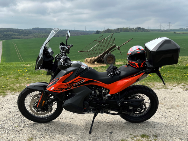 Ein KTM 1290 Super Adventure R Motorrad auf einem Schotterweg mit Helm und Gepäckstück daneben, umgeben von grünem Gras, hohen Bäumen und Strommästen unter einem bewölkten Himmel.