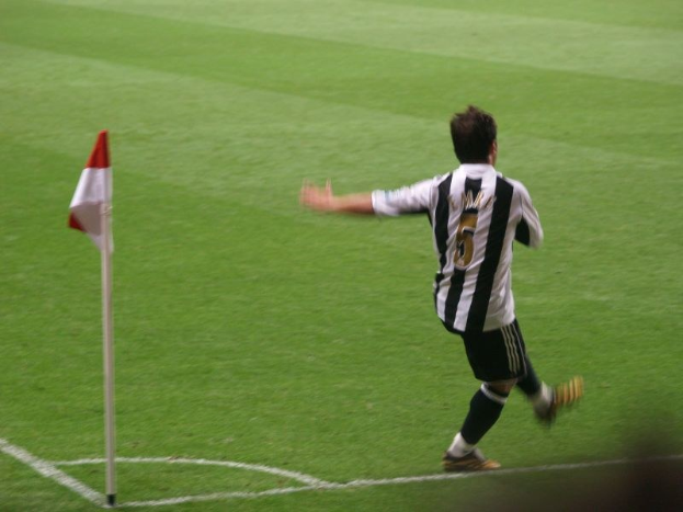 Ein Mann in einem schwarzen und weißen T-Shirt und schwarzen Shorts läuft auf einem Fußballfeld, feiert ein Tor mit erhobenen Armen, mit einer roten und weißen Fahne auf einem Pfahl links von ihm.