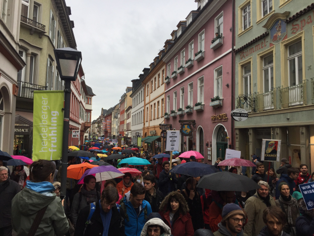 Große Gruppe von Menschen, die mit Schirmen die Straße entlanggehen, einige tragen Taschen und halten Schilder, während sie an einem Klimastreik in Deutschland teilnehmen.
