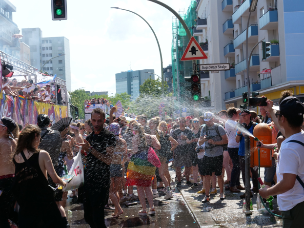 Menschen bei einer Christopher Street Day Parade bespritzen sich gegenseitig mit Wasser auf einer Stadtstraße, mit einem Banner, Gebäuden, Bäumen und Ampeln im Hintergrund.