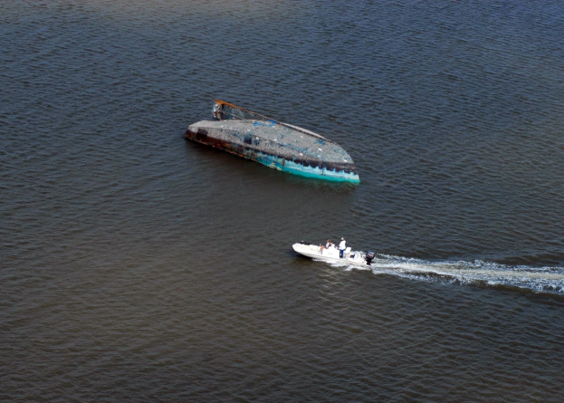 Ein kleines, teilweise untergetauchtes Wrackboot, das neben einem größeren Boot auf einem Fluss fährt, mit ein paar Menschen an Bord.