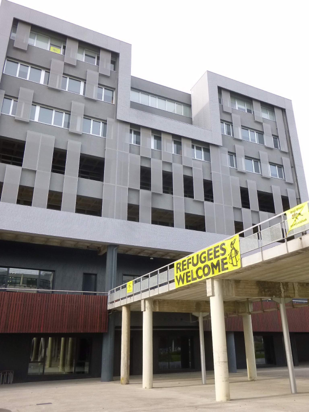 Ein großes Gebäude mit einer "Flüchtlinge Willkommen"-Schrift, umgeben von Säulen und einem Geländer, unter einem Himmel-Hintergrund.