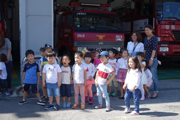 Gruppe von Kindern vor einem Feuerwehrfahrzeug an einer Feuerwache stehend, einige tragen Mützen, mit zusätzlichen Feuerwehrfahrzeugen im Hintergrund.