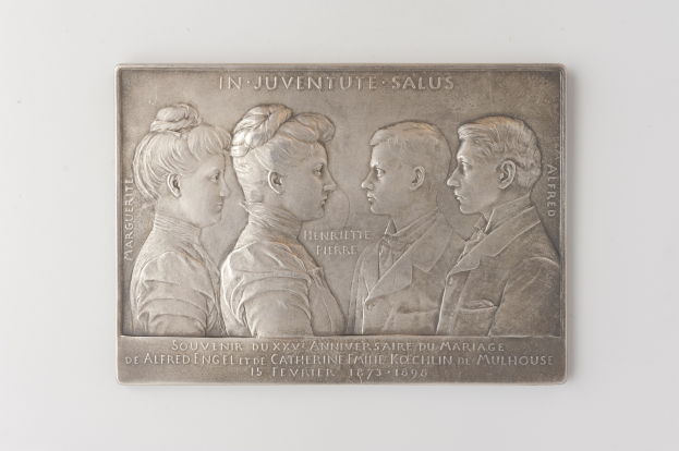 Silberne Plakette mit einem eingravierten Bild von drei Menschen und einem Gedenktext auf einem weißen Hintergrund.