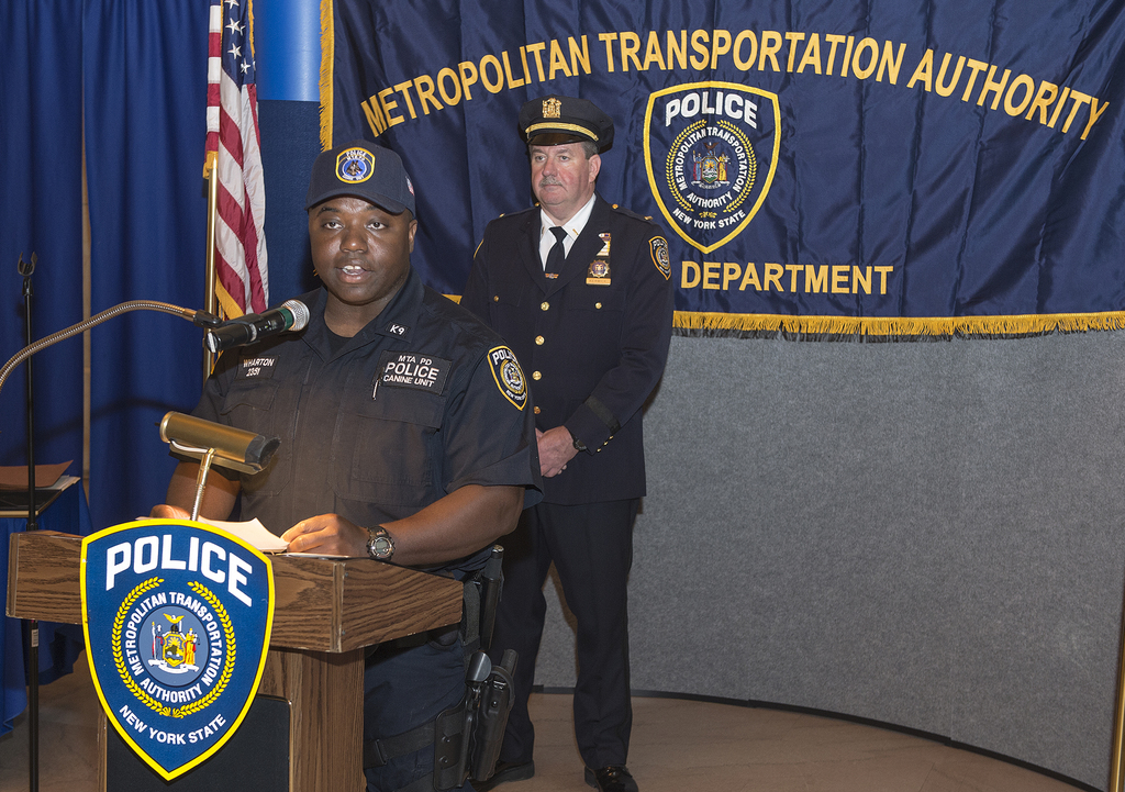 Zwei Polizeibeamte stehen vor einem Rednerpult, einer hält Papiere und spricht in ein Mikrofon, mit einer Tafel, Flagge und einem Tisch in der Nähe und einer Tafel mit Symbolen und Text im Hintergrund.