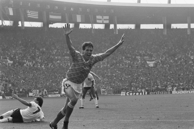 Schwarzes und weißes Foto eines Fußballspielers, der nach einem Tor in einem Stadion feiert, umgeben von Mannschaftskameraden und Mitarbeitern, mit Stadion-Elementen im Hintergrund.