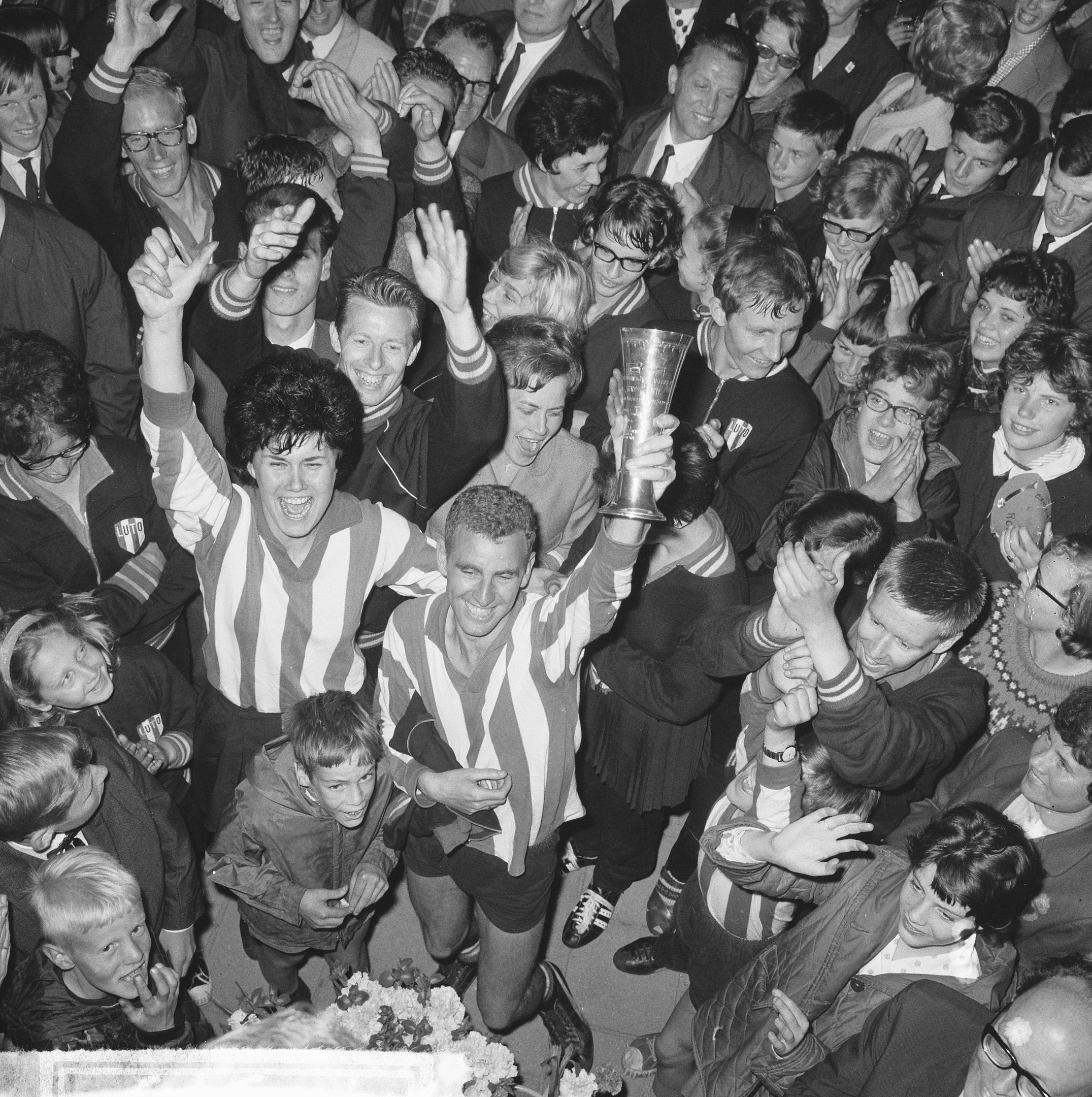 Schwarzes Bild einer Gruppe von Menschen, die mit einem Pokal feiern, eine Person in der Mitte hält die Tasse, Blumen unten, umgeben von einer Menge.