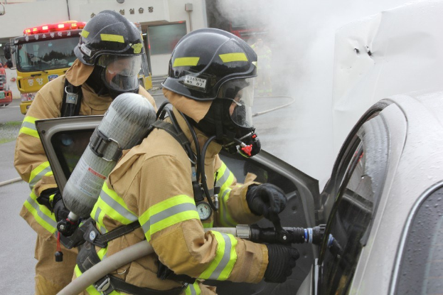 Zwei Feuerwehrleute in Schutzausrüstung verwenden einen Schlauch, um ein brennendes Auto zu löschen, mit Rauch sichtbar, während Fahrzeuge und ein Gebäude im Hintergrund sind.