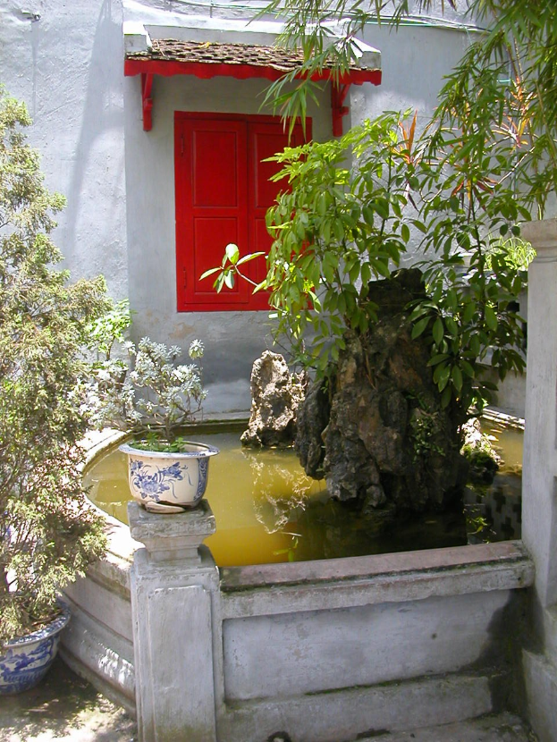 Ein kleiner Hof mit einer roten Tür, umgeben von Bäumen, Pflanzen, Felsen, einer Wand und einem Topf mit einer Pflanze.